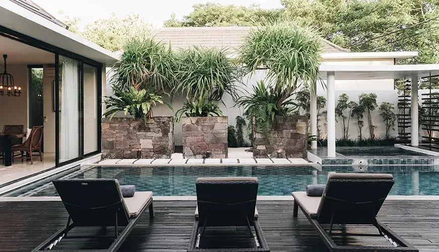 Ambergris Villa, Nusa Dua, Bali — three empty rattan lounge chairs facing swimming pool on wooden deck surrounded by tropical plants | The Luxury Bali