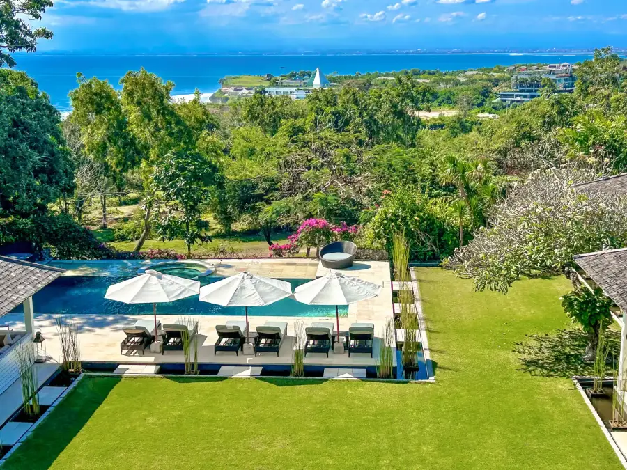 A ten-bedroom villa in Uluwatu, Bali: the pool, white umbrellas and lawn, with the coast and sea beyond the trees